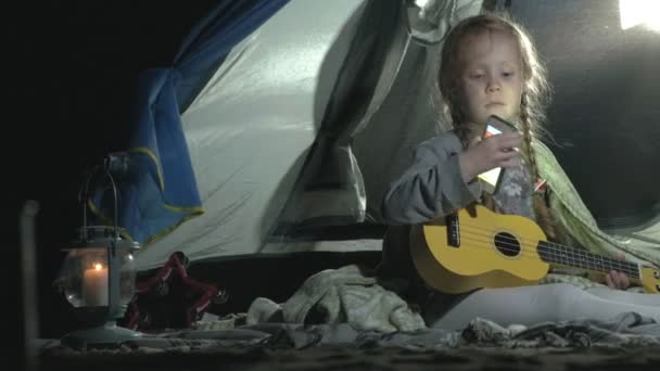 Krásná dívka si hraje na ukulele na břehu řeky poblíž turistické stany — Stock video