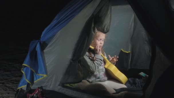 Uma menina bonita joga em um ukulele na margem do rio perto de uma tenda turística — Vídeo de Stock