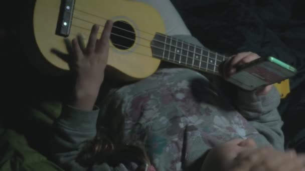 Una hermosa chica y mujer juega en un ukelele en la orilla del río cerca de una tienda de campaña turística — Vídeos de Stock