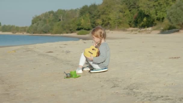 Uma menina bonita joga em um ukulele na margem do rio perto de uma tenda turística — Vídeo de Stock