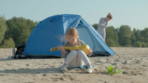 Een mooi meisje speelt op een ukulele aan de rivieroever in de buurt van een toeristische tent — Stockvideo