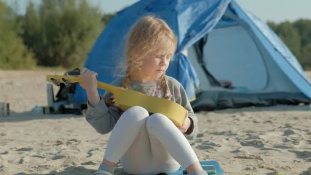 Een mooi meisje speelt op een ukulele aan de rivieroever in de buurt van een toeristische tent — Stockvideo