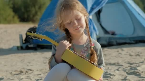 Uma menina bonita joga em um ukulele na margem do rio perto de uma tenda turística — Vídeo de Stock