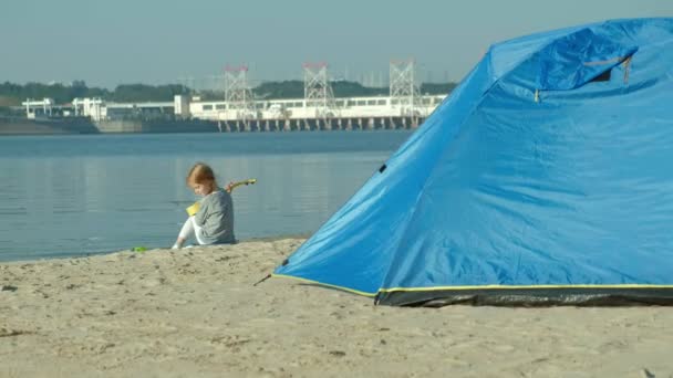 En vacker flicka spelar på en ukulele på flodstranden nära turist tält — Stockvideo