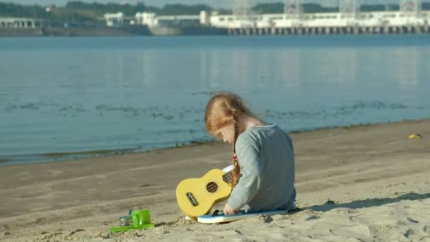 Krásná dívka si hraje na ukulele na břehu řeky poblíž turistické stany — Stock video