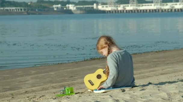 Una bella ragazza gioca su un ukulele sulla riva del fiume vicino a una tenda turistica — Video Stock