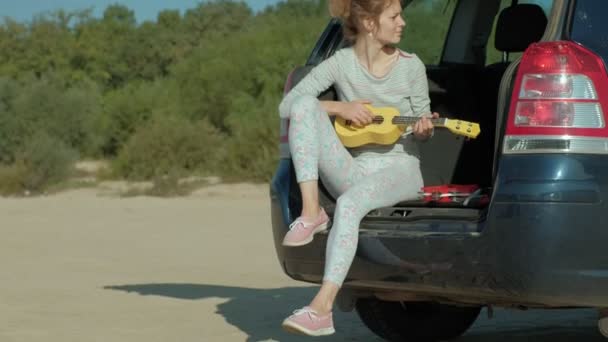 Kvinna som spelar på en ukulele i en öppen bål bil nära havet — Stockvideo