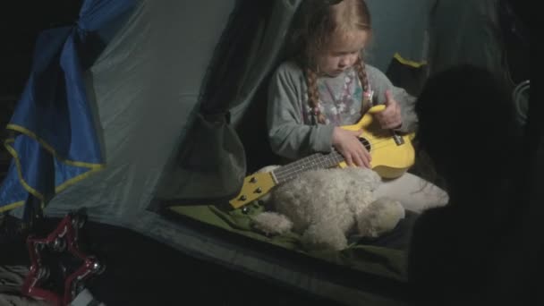 A beautiful girl plays on a ukulele on the river bank near a tourist tent — Stock Video