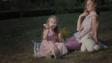 Mutlu anne ve kızı sabun köpüğü şişirmek. Aile içinde sıcak bir akşam günbatımında pikniğe üzerinde bir şehir parkı.