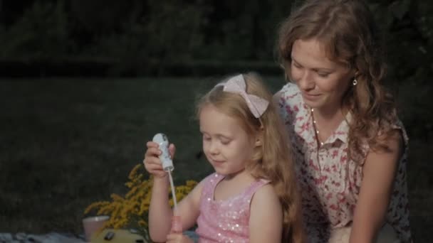 Une mère et une fille heureuses gonflent les bulles de savon. Famille dans un parc municipal lors d'un pique-nique lors d'une soirée chaude au coucher du soleil . — Video