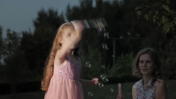 Une mère et une fille heureuses gonflent les bulles de savon. Famille dans un parc municipal lors d'un pique-nique lors d'une soirée chaude au coucher du soleil . — Video