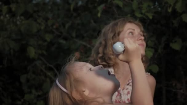 Une mère et une fille heureuses gonflent les bulles de savon. Famille dans un parc municipal lors d'un pique-nique lors d'une soirée chaude au coucher du soleil . — Video