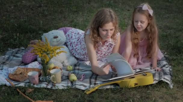 En glad mor och dotter Visa foton i ett album som en bok. Familjen i en park på en picknick på en varm kväll vid solnedgången. — Stockvideo