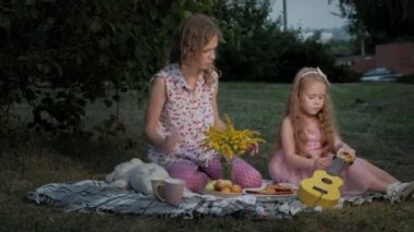 Mutlu anne ve kızı ukulele üzerinde oynuyorlar. Aile içinde sıcak bir akşam günbatımında pikniğe üzerinde bir şehir parkı.