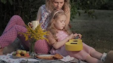 Mutlu anne ve kızı ukulele üzerinde oynuyorlar. Aile içinde sıcak bir akşam günbatımında pikniğe üzerinde bir şehir parkı.