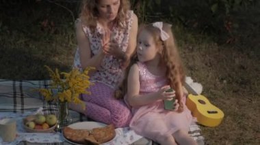 Mutlu anne ve kızı ukulele üzerinde oynuyorlar. Aile içinde sıcak bir akşam günbatımında pikniğe üzerinde bir şehir parkı.