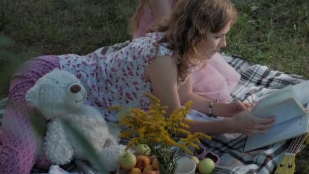 Una madre y una hija felices ven fotos en un álbum leen un libro. Familia en un parque de la ciudad en un picnic en una cálida noche al atardecer . — Vídeos de Stock