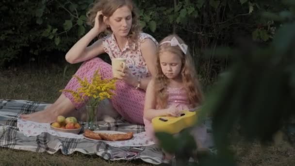 En glad mor och dotter spelar på ukulele. Familjen i en park på en picknick på en varm kväll vid solnedgången. — Stockvideo