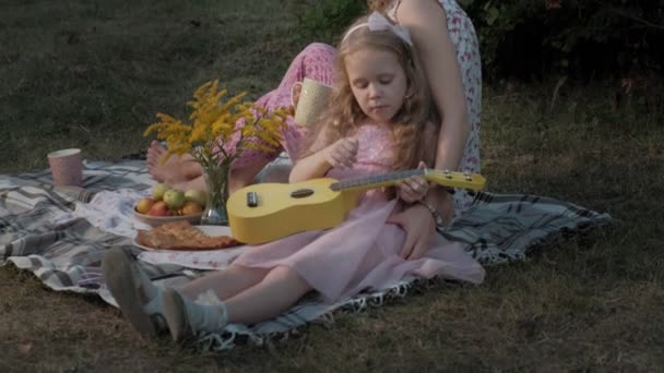 Eine glückliche Mutter und ihre Tochter spielen auf der Ukulele. Familie im Stadtpark bei einem Picknick an einem warmen Abend bei Sonnenuntergang. — Stockvideo