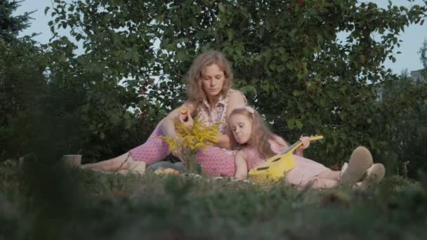 Uma mãe e uma filha felizes estão a brincar no ukulele. Família em um parque da cidade em um piquenique em uma noite quente ao pôr do sol . — Vídeo de Stock