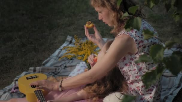 Eine glückliche Mutter und ihre Tochter spielen auf der Ukulele. Familie im Stadtpark bei einem Picknick an einem warmen Abend bei Sonnenuntergang. — Stockvideo