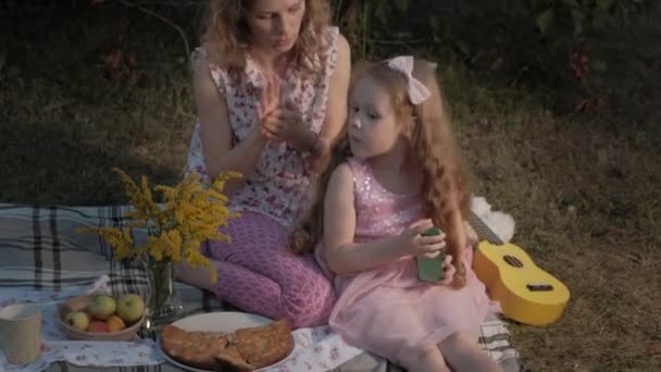 Eine glückliche Mutter und ihre Tochter spielen auf der Ukulele. Familie im Stadtpark bei einem Picknick an einem warmen Abend bei Sonnenuntergang. — Stockvideo