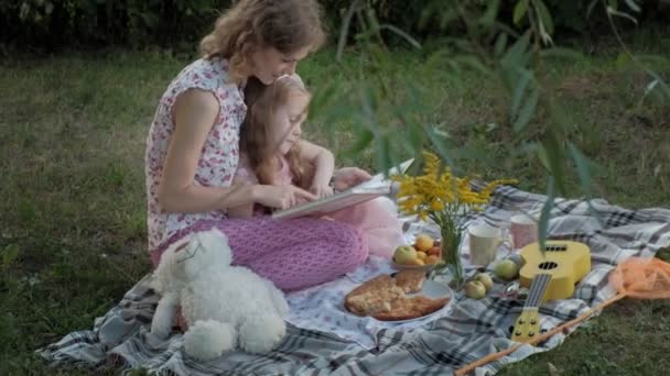 Een gelukkige moeder en dochter bekijken foto's in een album, een boek lezen. Familie in een stadspark op een picknick op een warme avond bij zonsondergang. — Stockvideo