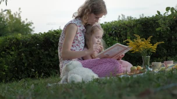 Uma mãe e uma filha felizes veem fotos em um álbum lêem um livro. Família em um parque da cidade em um piquenique em uma noite quente ao pôr do sol . — Vídeo de Stock