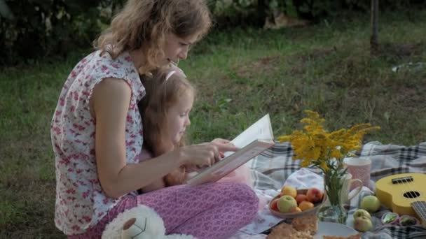 Uma mãe e uma filha felizes veem fotos em um álbum lêem um livro. Família em um parque da cidade em um piquenique em uma noite quente ao pôr do sol . — Vídeo de Stock