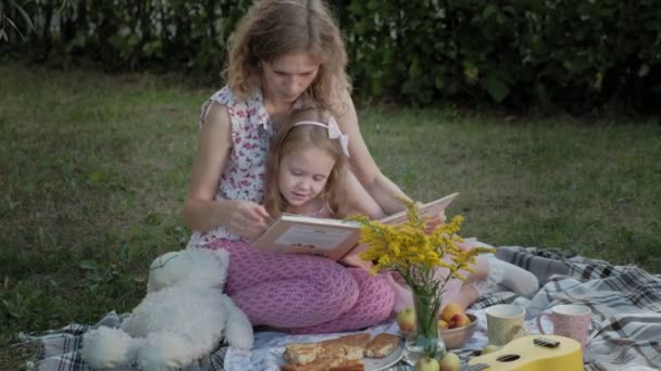 Een gelukkige moeder en dochter bekijken foto's in een album, een boek lezen. Familie in een stadspark op een picknick op een warme avond bij zonsondergang. — Stockvideo