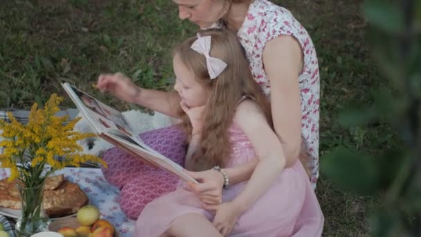 Mutlu anne ve kızı bir kitap okumak bir albümdeki fotoğrafları. Aile içinde sıcak bir akşam günbatımında pikniğe üzerinde bir şehir parkı. — Stok video