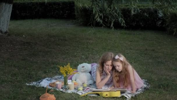 一个快乐的母亲和女儿在相册中查看照片读了一本书。在一个城市公园的家庭在一个温暖的傍晚在日落野餐. — 图库视频影像