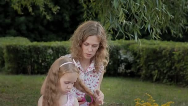 Eine glückliche Mutter kämmt ihren Töchtern die Haare. Familie im Stadtpark bei einem Picknick an einem warmen Abend bei Sonnenuntergang. — Stockvideo