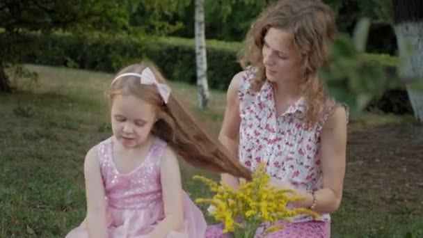 Una madre feliz peina el pelo de sus hijas. Familia en un parque de la ciudad en un picnic en una cálida noche al atardecer . — Vídeo de stock