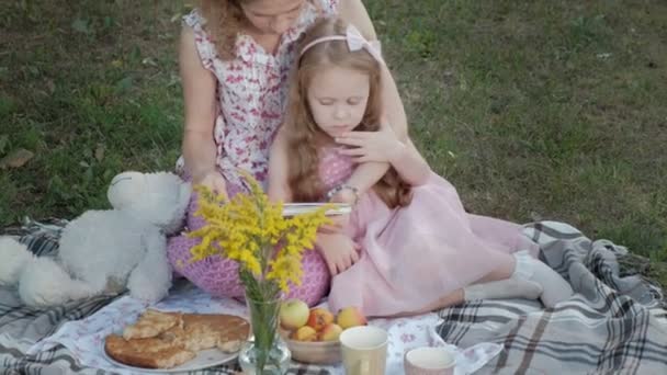 Eine glückliche mutter und tochter sehen fotos in einem album lesen ein buch. Familie im Stadtpark bei einem Picknick an einem warmen Abend bei Sonnenuntergang. — Stockvideo