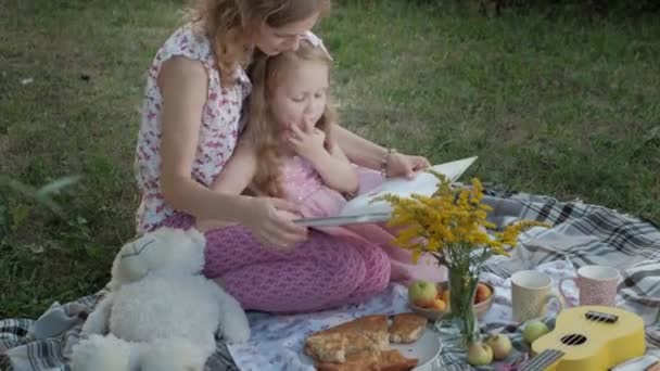 En glad mor och dotter Visa foton i ett album som en bok. Familjen i en park på en picknick på en varm kväll vid solnedgången. — Stockvideo