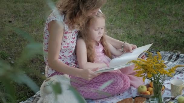 Щаслива мати і дочка переглядають фотографії в альбомі, читаючи книгу. Сім'я в міському парку на пікніку в теплий вечір на заході сонця . — стокове відео
