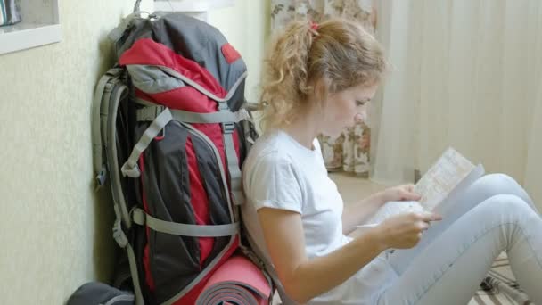 Turista mujer recoge cosas en una mochila en la cocina de la casa y se prepara para el viaje mira el mapa de papel — Vídeo de stock