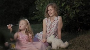 Mutlu anne ve kızı sabun köpüğü şişirmek. Aile içinde sıcak bir akşam günbatımında pikniğe üzerinde bir şehir parkı.