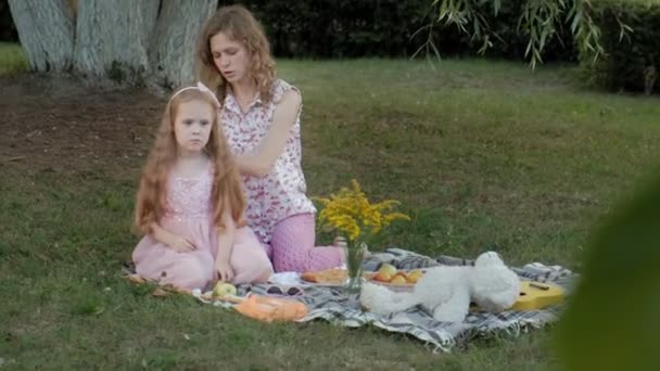 Счастливая мать расчесывает своих дочерей. Семья в городском парке на пикнике в теплый вечер на закате . — стоковое видео