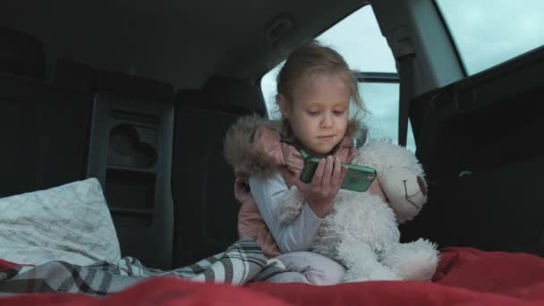 Menina bonita sentada em um tronco aberto de um carro na margem do rio do mar usa um smartphone — Vídeo de Stock