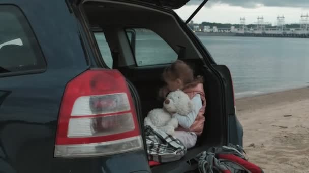 Vacker liten flicka sitter i en öppen bakluckan på en bil på flodstranden av havet använder en smartphone — Stockvideo