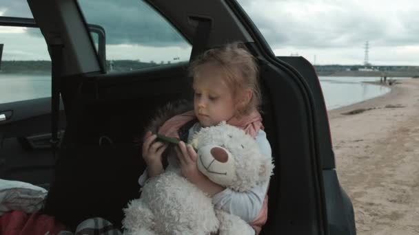 Красива маленька дівчинка сидить у відкритому багажнику автомобіля на березі моря використовує смартфон — стокове відео
