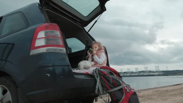 Hermosa niña sentada en un baúl abierto de un coche en la orilla del río del mar utiliza un teléfono inteligente — Vídeo de stock