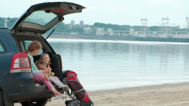 Mulher nova bonita e sua filha pequena estão sentados no tronco aberto de um carro na margem de um rio do mar comendo frutas e bebendo chá de uma garrafa térmica — Vídeo de Stock