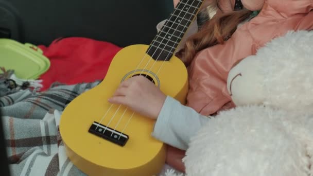 Menina bonita sentada em um tronco aberto de um carro na margem do rio do mar jogando em um ukulele amarelo — Vídeo de Stock