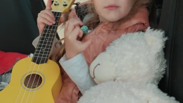 Красивая маленькая девочка, сидящая в открытом багажнике машины на берегу моря и играющая на желтой укулеле — стоковое видео
