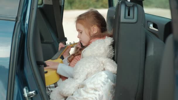 Όμορφο κοριτσάκι που κάθεται σε ένα ανοιχτό πορτμπαγκάζ του αυτοκινήτου στην όχθη του ποταμού στη θάλασσα παίζει σε ένα κίτρινο γιουκαλίλι — Αρχείο Βίντεο