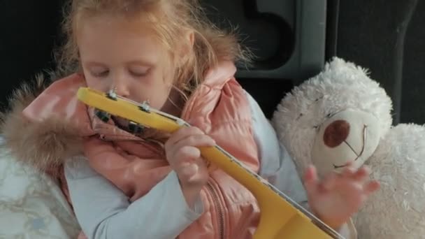 Hermosa niña sentada en un baúl abierto de un coche en la orilla del río del mar jugando en un ukelele amarillo — Vídeo de stock