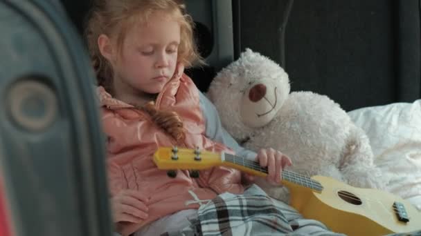 Belle petite fille assise dans un coffre ouvert d'une voiture sur la rive de la mer jouant sur un ukulélé jaune — Video